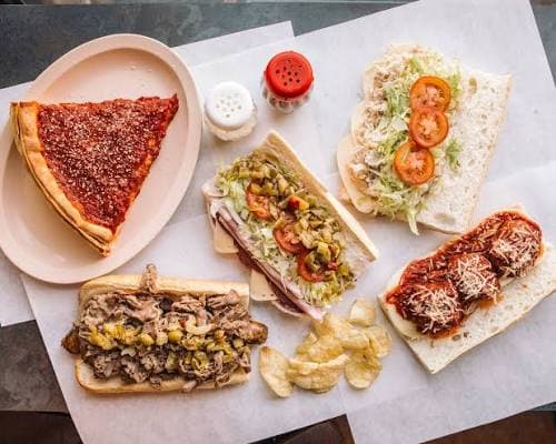Fontano’s spread on butcher paper — a slice of deep dish, an Italian beef, a giardiniera sub, a chicken salad sandwich, and a meatball sub with chips